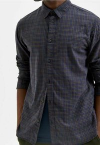 Camicia a righe blu con bottoni e colletto. Il tessuto è intrecciato, presenta un motivo a scacchi in varie tonalità ed è caratterizzata da una vestibilità ampia con maniche lunghe.