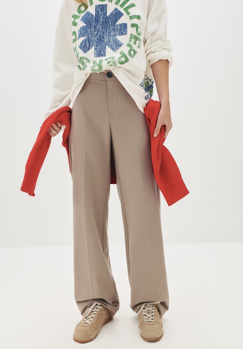 Personne portant un pantalon beige à jambes larges, des baskets beiges, un sweat-shirt blanc avec des dessins bleus et verts, tenant un pull rouge sur les côtés.