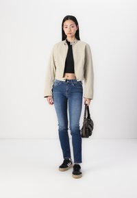 Beżowa kurtka cropped, czarny dopasowany top, niebieskie skinny jeans i czarne trampki w białe paski. Trzyma brązową torebkę. Prosty design, gładka tekstura.