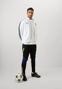 Balta sporta jaka ar melniem akcentiem un logotipu, saskaņota ar melnām un zilām biksēm. Melni futbola apavi un futbola bumba. Vienkāršs fons.