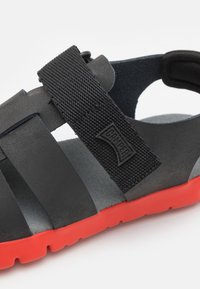 Musta sandaali, jossa on teksturoitu kangashihnat ja punainen kumipohja. Hihnassa on Camper-logot, suunniteltu mukavuutta ja monipuolisuutta varten.