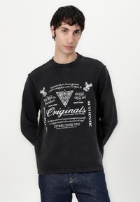 Sorte sweatshirt med rund halsudskæring, der har trykt hvid tekst og grafik. Lange ærmer, blød tekstur og afslappet pasform. Båret med blå jeans.