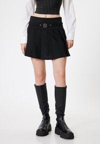 Schwarzer plissierter Minirock mit breitem Bund und Gürtel, kombiniert mit schwarzen knielangen Stiefeln mit robuster Sohle und Reißverschlussdetails.