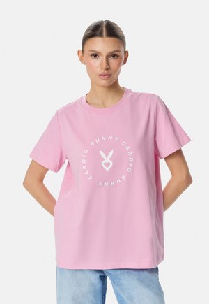 Naine kannab heleroosat T-särki valge ümmarguse "Cardio Bunny" logoga ja hele siniseid teksaseid, seisab käed selja taga.