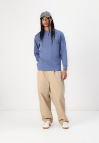 Chemise bleu clair à manches longues avec logo, pantalon large beige, et casquette verte. Les chaussures ont une base blanche avec des rayures rouges. Design minimaliste.