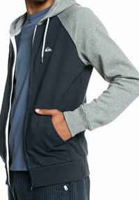 Navy and grey zip-up hoodie with a drawstring hood, contrasting sleeves, a small brand logo, and side pockets. Made from soft, blended fabric.