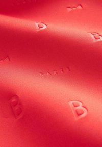 Koronkowy materiał w kolorze koralu, z wytłoczonymi literami "B" oraz wzorem w kokardy, o gładkiej, lekko błyszczącej teksturze i subtelnym wzorze.
