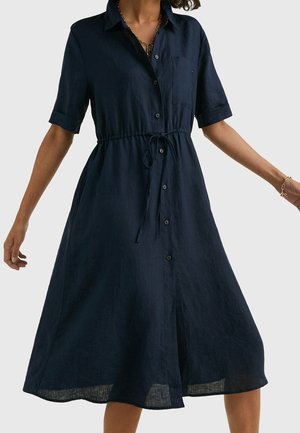 Frau trägt ein marineblaues Hemdkleid mit kurzen Ärmeln und gebundener Taille, steht mit leicht erhobenen Armen.