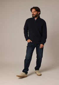 Un pull marine avec un tissu tricoté texturé et un col zippé mi-haut, associé à un jean sombre et des chaussures de couleur claire.