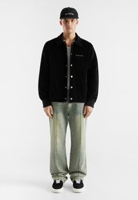 Black corduroy jacket with snap buttons, gray denim trousers, and black sneakers with white laces. Black cap with logo on front.