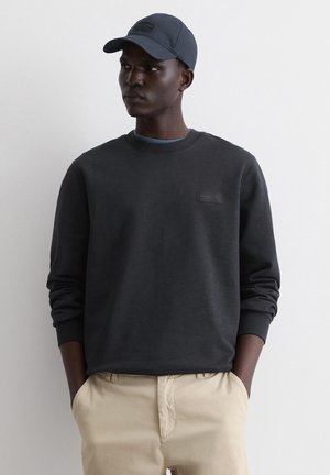 Dark gray sweatshirt with a round neck, long sleeves, and a small textured label. Paired with a matching cap and beige pants.