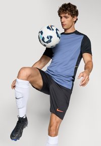 Blå og svart treningsgenser med korte ermer, kombinert med svarte shorts. Hvite sokker med et logo, svarte fotballsko og en flerfarget fotball.