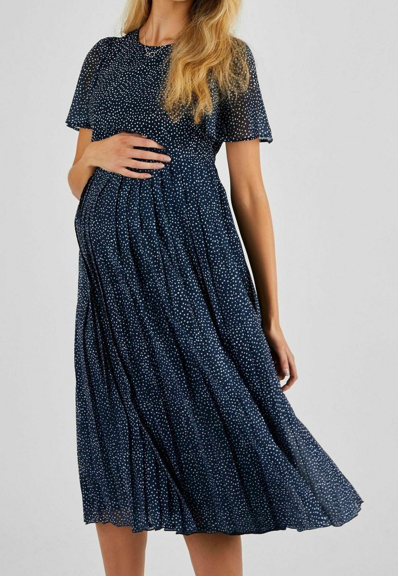 Femme enceinte portant une robe midi bleu marine à pois blancs, main posée sur son ventre, debout devant un fond uni.