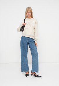 Lichtbeige sweatshirt met bloemen borduursel, ruime lichtblauwe spijkerbroek, en zwarte geschenkte schoenen. Model houdt een kleine zwarte handtas vast.