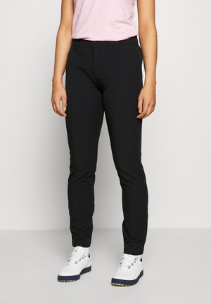 Vrouw draagt strakke zwarte broek, lichtroze mouwloze top en witte sneakers, staand tegen een effen lichte achtergrond.