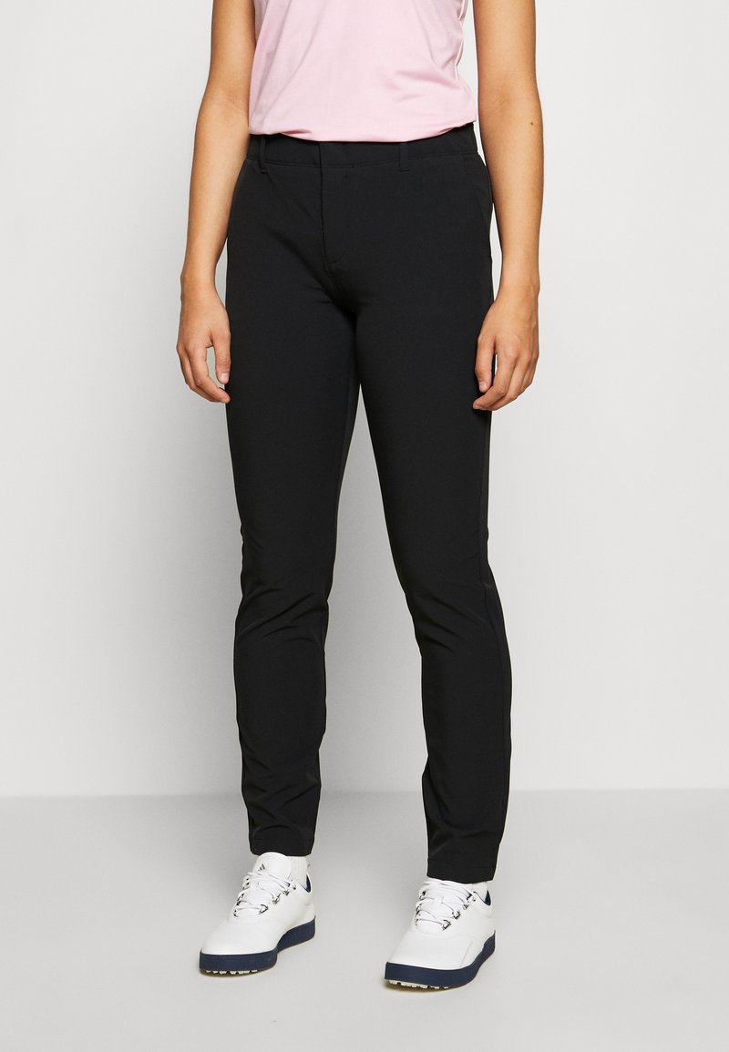 Femme portant un pantalon noir ajusté, un haut sans manches rose clair et des baskets blanches, debout devant un fond clair uni.