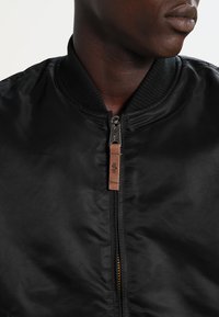 Veste bomber noire avec une finition brillante, dotée d'un col côtelé, d'une fermeture éclair et d'une étiquette en cuir avec un détail de logo. Texture lisse.