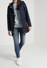 Marinblå regnjacka med polka prickar och huva, med stora vita prickar, knappar fram och dragsko. Kombinerad med ljusblå tröja och jeans.