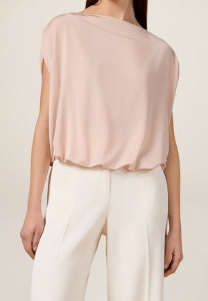 Frau trägt eine hellrosa, lockere ärmellose Bluse, die in hoch taillierte weiße Hose mit weitem Bein gesteckt ist.