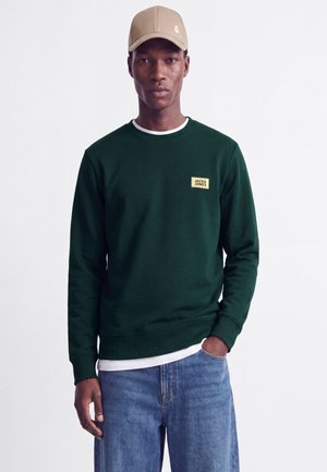 Homme portant un sweat-shirt vert foncé Jack & Jones, une casquette beige clair, un sous-maillot blanc et un jean bleu, debout sur un fond uni.