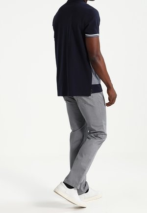 Mand iført en marineblå polo, lysegrå bukser og hvide sneakers, stående med det ene ben let bøjet mod en hvid baggrund.