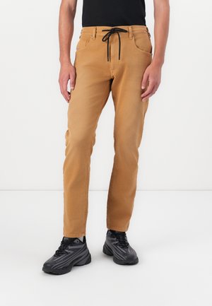 Mann trägt hellbraune, gerade geschnittene Hosen mit schwarzem Kordelzug und schwarzen Sportschuhen, steht vor einem weißen Hintergrund.