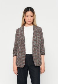 Blazer a quadri in tonalità di marrone e blu, con una vestibilità su misura, apertura frontale e maniche arrotolate. Indossato sopra un top bianco.
