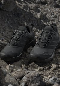 Paire de chaussures de randonnée Adidas Terrex noires avec des lacets réfléchissants posées sur un terrain rocheux en extérieur.