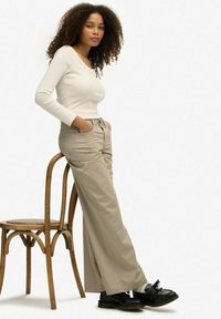 Vrouw met krullend haar leunend tegen een houten stoel, gekleed in een witte blouse met lange mouwen, beige wijde broek en zwarte loafers.