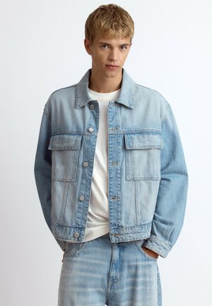 Jeune homme portant une veste en jean bleu clair et un jean assorti avec un t-shirt blanc, debout les mains dans les poches devant un fond uni.