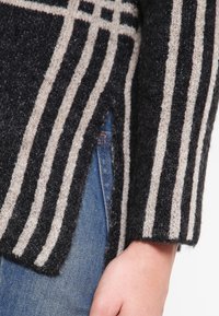 Zbliżenie na rękaw swetra w czarne i beżowe paski z dzianiny oraz boczny rozcięcie, noszonego na niebieskich dżinsach, z widoczną dłonią.