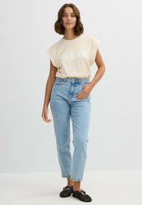Camiseta beige de manga corta con texto gráfico blanco, combinada con unos jeans de tiro alto azul claro y zapatos planos de malla negros. Diseño sencillo.