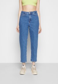 Džínové kalhoty s vysokým pasem v modré barvě, rovného střihu, s knoflíkovým zapínáním a designem se pěti kapsami. Nosené s béžovými teniskami.