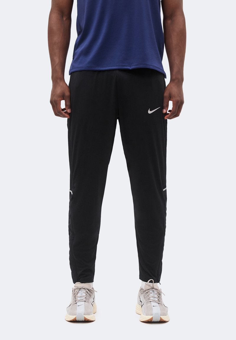 Άνδρας που φοράει μαύρο αθλητικό παντελόνι Nike και ανοιχτογκρι παπούτσια για τρέξιμο, στέκεται με τα χέρια χαλαρά στα πλευρά σε απλό φόντο.