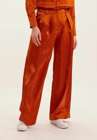 Pantalons larges en satin orange à taille haute, avec des plis avant et une ceinture assortie. Stylés avec des baskets blanches.