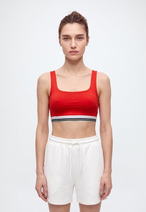 Une femme se tient devant un fond blanc uni, vêtue d’un crop top rouge bordé de rayures et d’un short blanc à cordon de serrage.