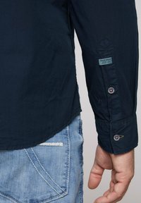 Camicia a maniche lunghe blu navy con polsini abbottonati, con un'etichetta con il testo "CAMP DAVID". Jeans azzurri visibili con cuciture bianche sulla tasca.