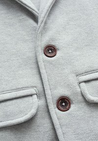 Ljust grått texturerat tyg med två bruna knappar märkta "MNG KIDS" och två fickor med lock.