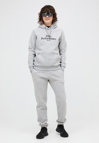 Grå bomulds-hættetrøje med frontlomme og sort logo; matchet med grå sweatpants og sorte atletiske sko. Modellen har mørke solbriller på.