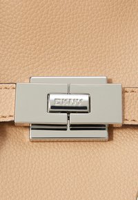 Bežová kožená kabelka s texturovaným povrchem a stříbrným kovovým zapínáním s logem "DKNY". Ozdobné prošívání podél popruhu.