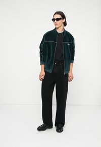 Zelená zamatová bomber bunda s bielym lemovaním, čierne tričko, široké čierne džínsy a čierne topánky; model nosí tmavé slnečné okuliare.