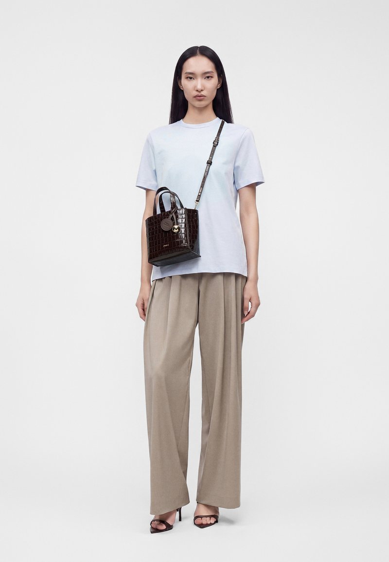 Femme portant un t-shirt bleu clair et un pantalon large beige, portant un sac à motif crocodile marron foncé avec une bandoulière, debout sur fond blanc.