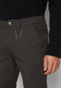 Homme portant un pull noir et un pantalon foncé avec un mousqueton argenté accroché à la passants de ceinture au niveau de la taille.