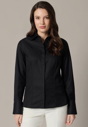 Kobieta z długimi brązowymi włosami, ubrana w dopasowaną czarną koszulę zapinaną na guziki i białe spodnie, stojąca na jednolitym beżowym tle.