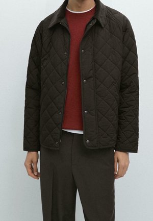 Mężczyzna w ciemnobrązowej pikowanej kurtce na rdzawym swetrze i brązowych spodniach na jednolitym jasnym tle.