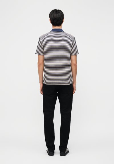 Άνδρας που κοιτάζει προς τα πίσω, φοράει μπλε σκούρα και λευκή ριγέ κοντομάνικη πόλο μπλούζα, μαύρο παντελόνι και μαύρα παπούτσια, με φόντο ένα απλό λευκό φόντο.