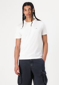 Witte katoenen T-shirt met korte mouwen, ronde halslijn en klein geborduurd logo op de borst, gedragen met donkere cargoshorts.