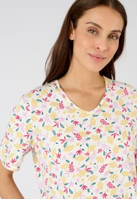 Camisa de manga corta con cuello en V, hecha de una tela ligera, con un patrón floral que presenta elementos en rosa, amarillo y verde azulado sobre un fondo crema.