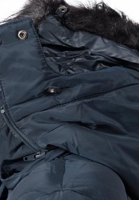 Veste d'hiver bleu foncé avec une capuche doublée de fausse fourrure noire, fermeture éclair et boutons-pression visibles au niveau du col.
