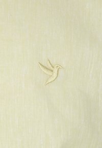 Heled kollane riie näitab vasakule poolse väikese tikandiga linnu embleemi, mis on veidi tumedamas kollases. Kude paistab pehme.